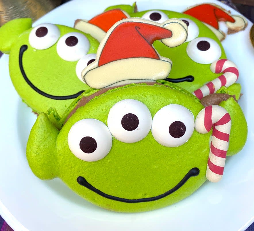 A macaron shaped like the head of an alien from Toy Story, with a santa hat and candy cane made of frosting by its side