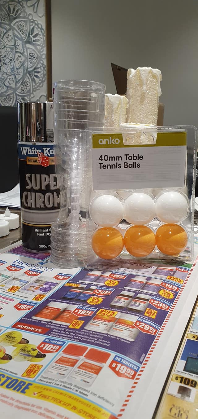 A pack of table tennis balls, silver spray paint and plastic wine glasses on top of a sheet of newspaper.
