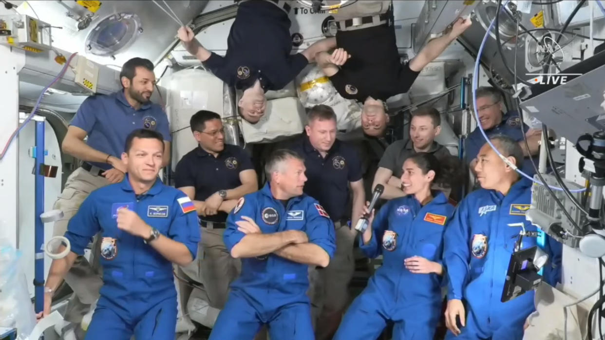  Eleven astronauts in different color uniforms gather on a space station, two float upside down. 