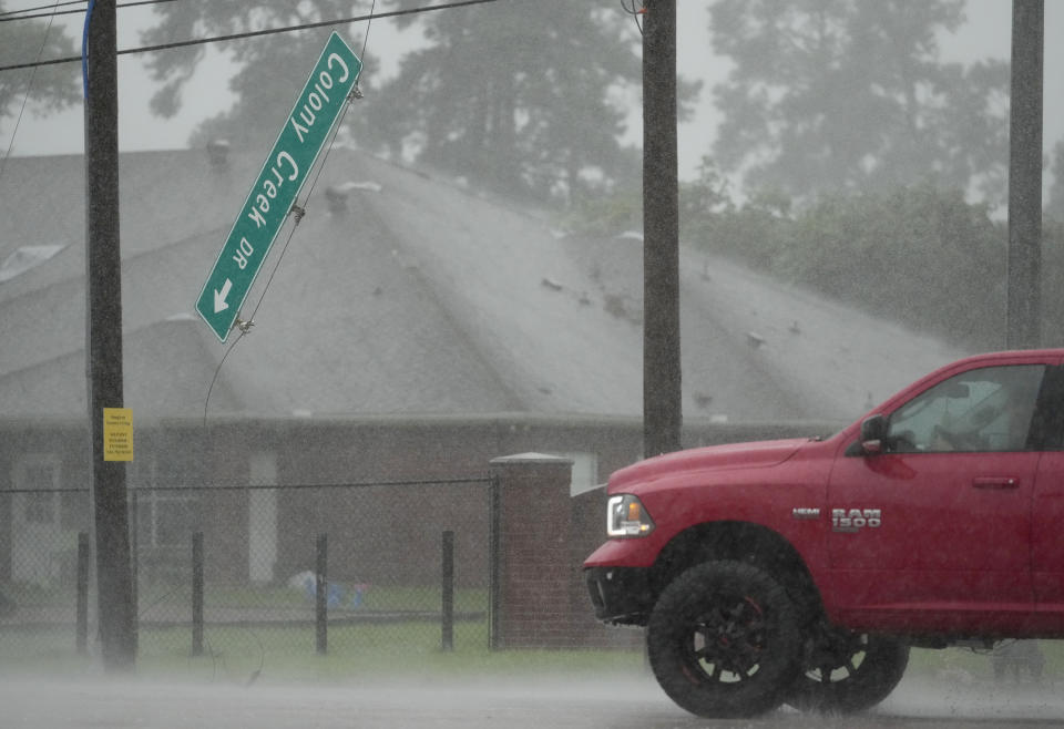 A red truck drives through heavy rain past a damaged "Cody Creek Rd" street sign and a building in the background. The weather is stormy with limited visibility