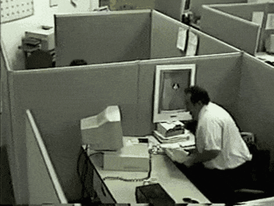 A man hitting his computer monitor off his desk with his keyboard
