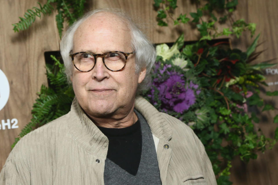 Chevy Chase, in a casual outfit with a light jacket and glasses, stands in front of a backdrop adorned with greenery and flowers