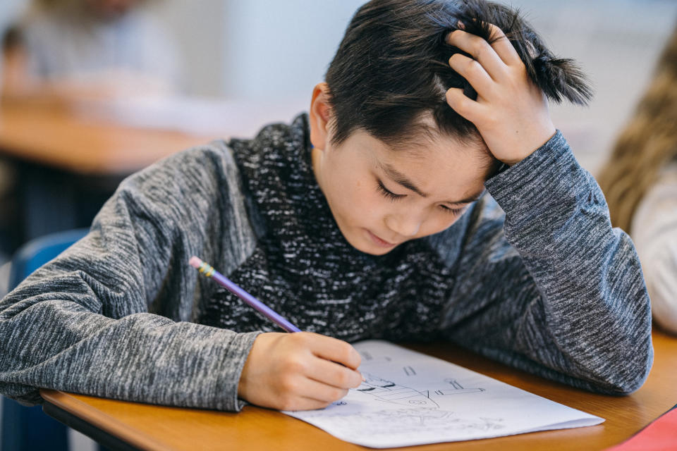 A young child concentrates while holding a pencil and scratching their head, working on schoolwork at a desk in a classroom