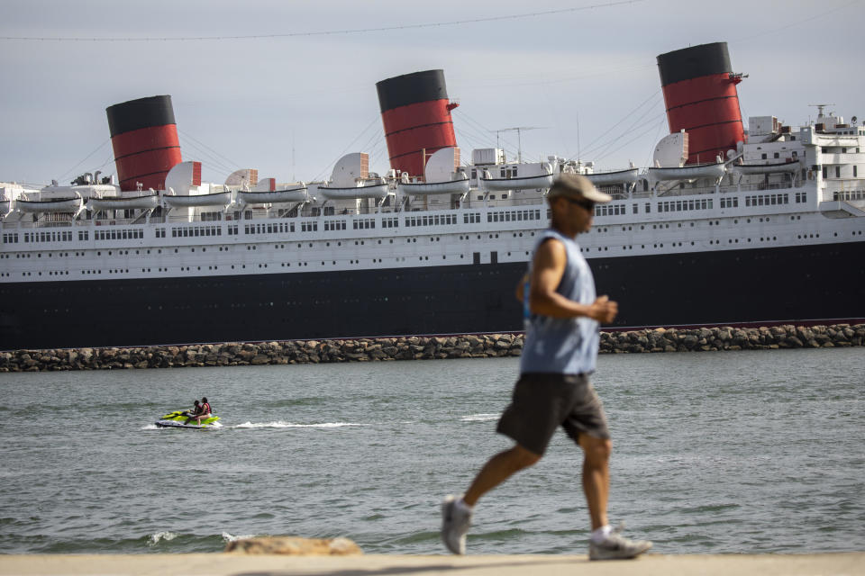 Two people on a jetski are seen in the water, while a runner passes by along Shoreline Aquatic Park, with the Queen Mary ship in the distance, docked in Long Beach, CA,