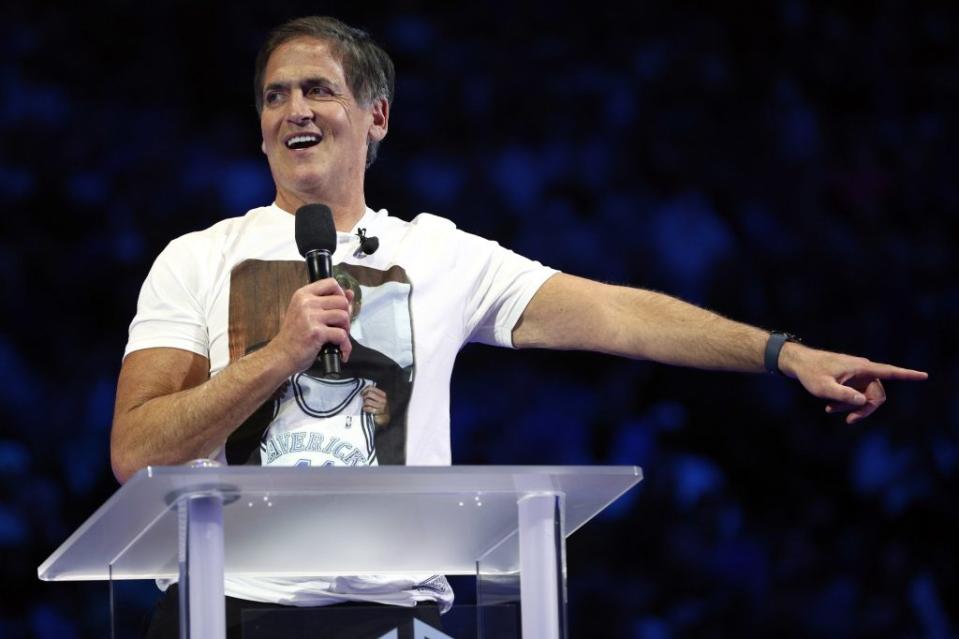 Mark Cuban stands at a podium smiling. He holds a mic with one hand and is pointing off-stage with the other.