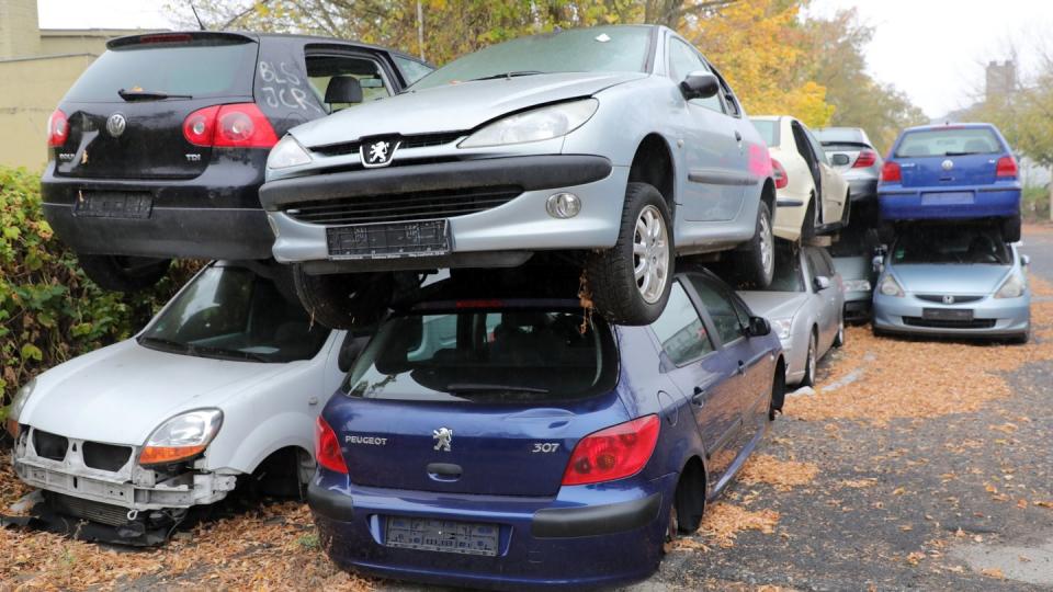 Vor dem Gelände eines Schrottplatzes mit Autopresse stapeln sich alte Autos, vorwiegend Dieselfahrzeuge.