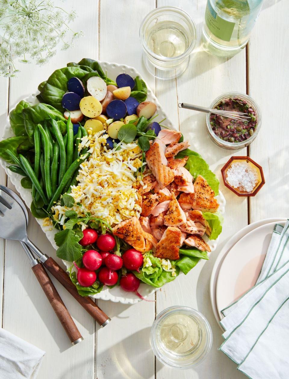 seared salmon watercress and potato salad with olive dressing on a large white serving plate with serving utensils and small bowls of toppings on the side