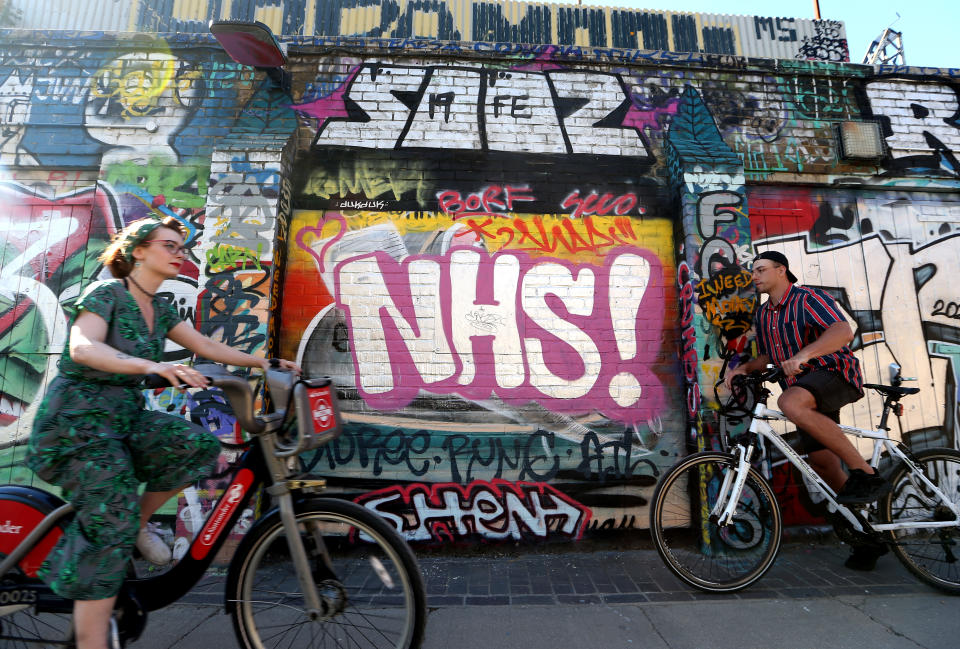 LONDON, ENGLAND  - MAY 31: Cyclists pass graffiti reading 'NHS'  in Hackney Wick on May 31, 2020 in London, England. The British government continues to ease the coronavirus lockdown by announcing schools will open to reception year pupils plus years one and six from June 1st. Open-air markets and car showrooms can also open from the same date.  (Photo by Alex Pantling/Getty Images)