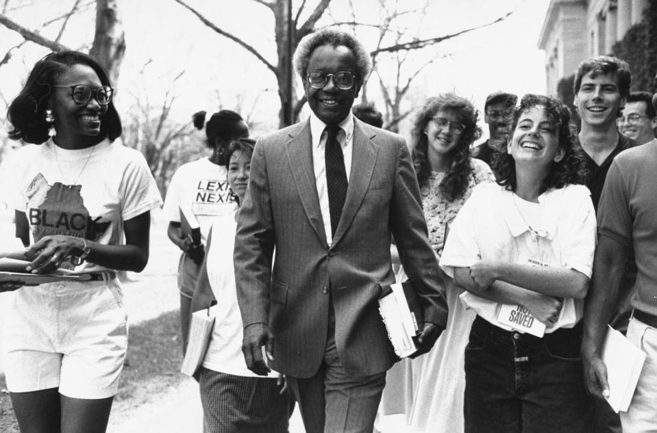 A black-and-white photo of a man wearing a suit walking with a group of students, each one carrying a book.