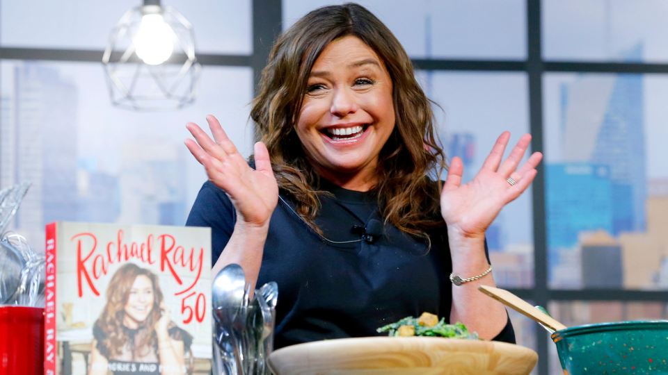 Rachael Ray smiling during a filmed appearance, with bowls of food and her cookbook on a table in front of her