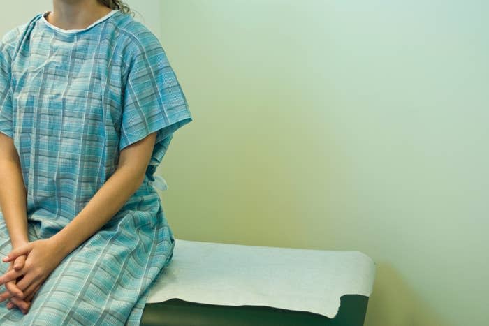 A person sitting on a medical examination table, wearing a hospital gown and with their hands folded in their lap