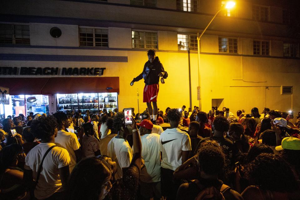 A man stands on a car as crowds defiantly gather in the street while a speaker blasts music an hour past curfew in Miami Beach, Fla., on Sunday, March 21, 2021. An 8 p.m. curfew has been extended in Miami Beach after law enforcement worked to contain unruly crowds of spring break tourists. (Daniel A. Varela/Miami Herald via AP)