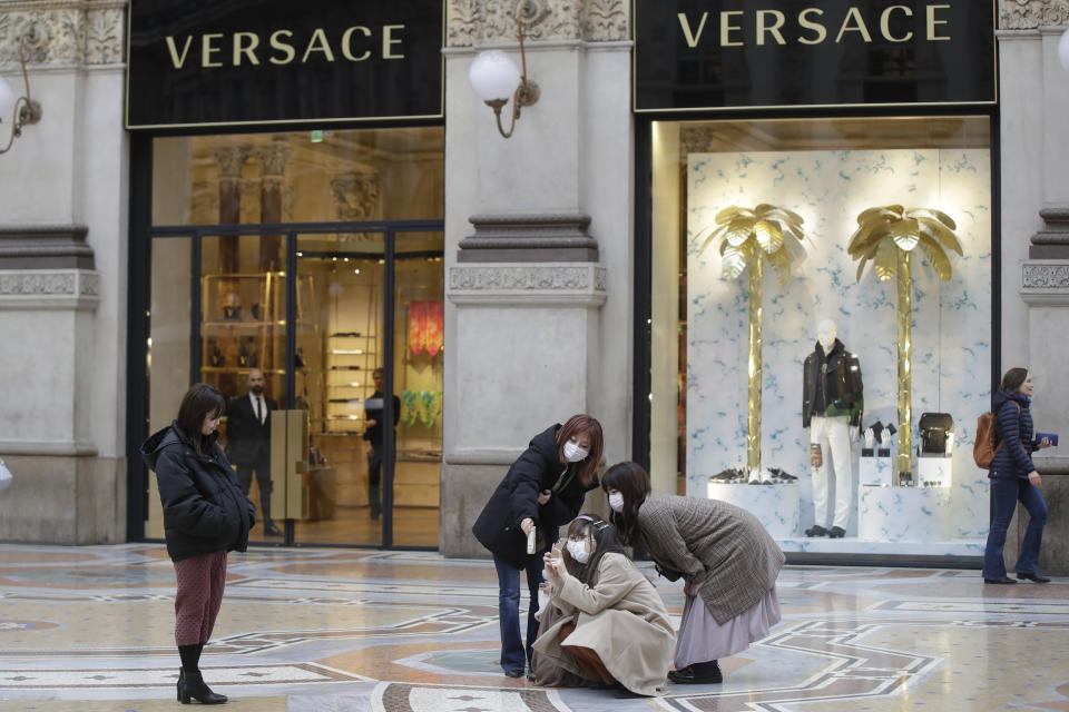 Tourists, wearing face masks, pose for a selfie in front of a Versace shop window in downtown Milan, Italy, Thursday, Feb. 27, 2020. In Europe, an expanding cluster in northern Italy is eyed as a source for transmissions. (AP Photo/Luca Bruno)