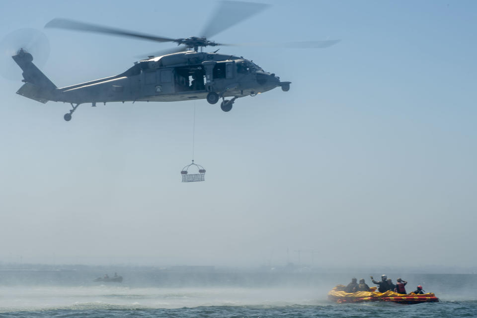 helicopter hovering over the ocean with a basket underneath. a raft on the ocean to the right holds a group of sailors