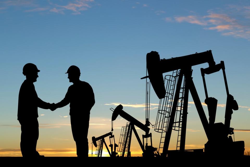 Two people shake hands while standing in front of oil drilling rigs.