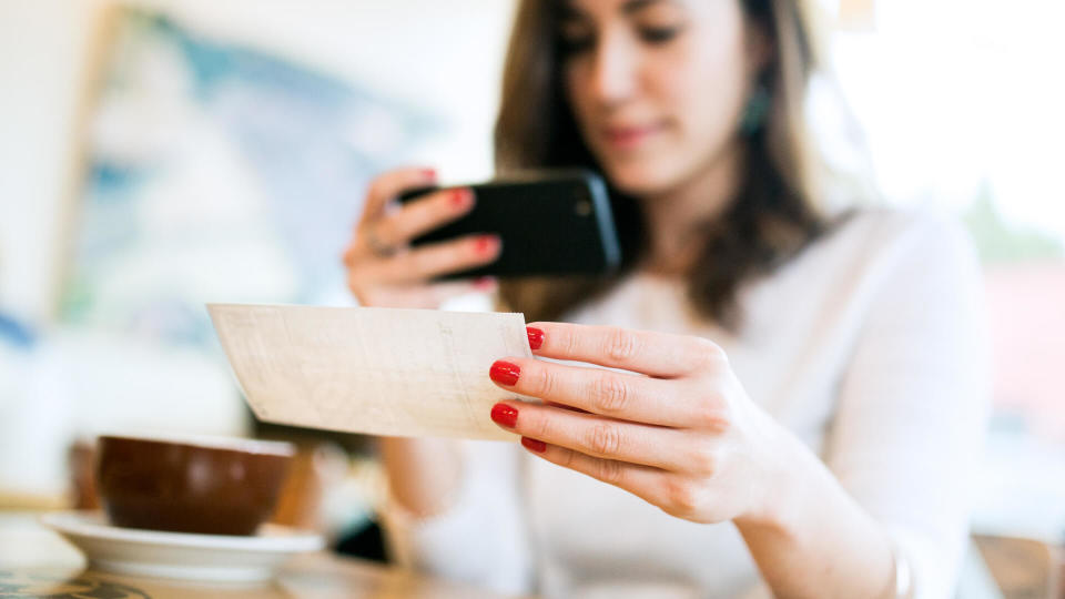 A smiling young woman takes a picture with her smart phone of a check or paycheck for digital electronic depositing, also known as 