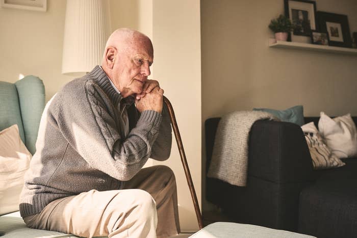 An elderly man in a sweater sits pensively on a couch, holding a cane, alone