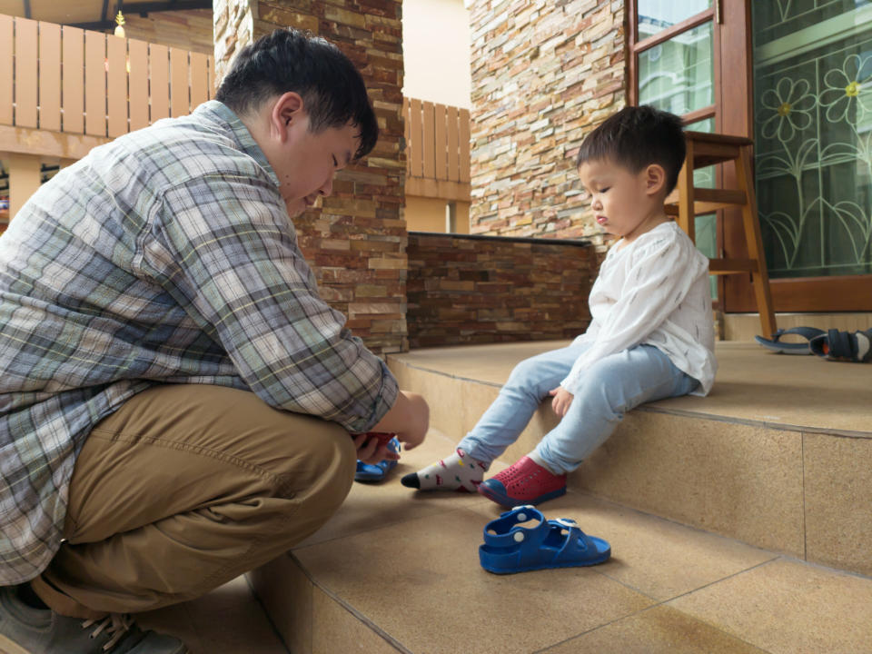 A man helps a child put on shoes while they both sit on stairs near a stone wall. The child, in a white shirt and light pants, has mismatched socks