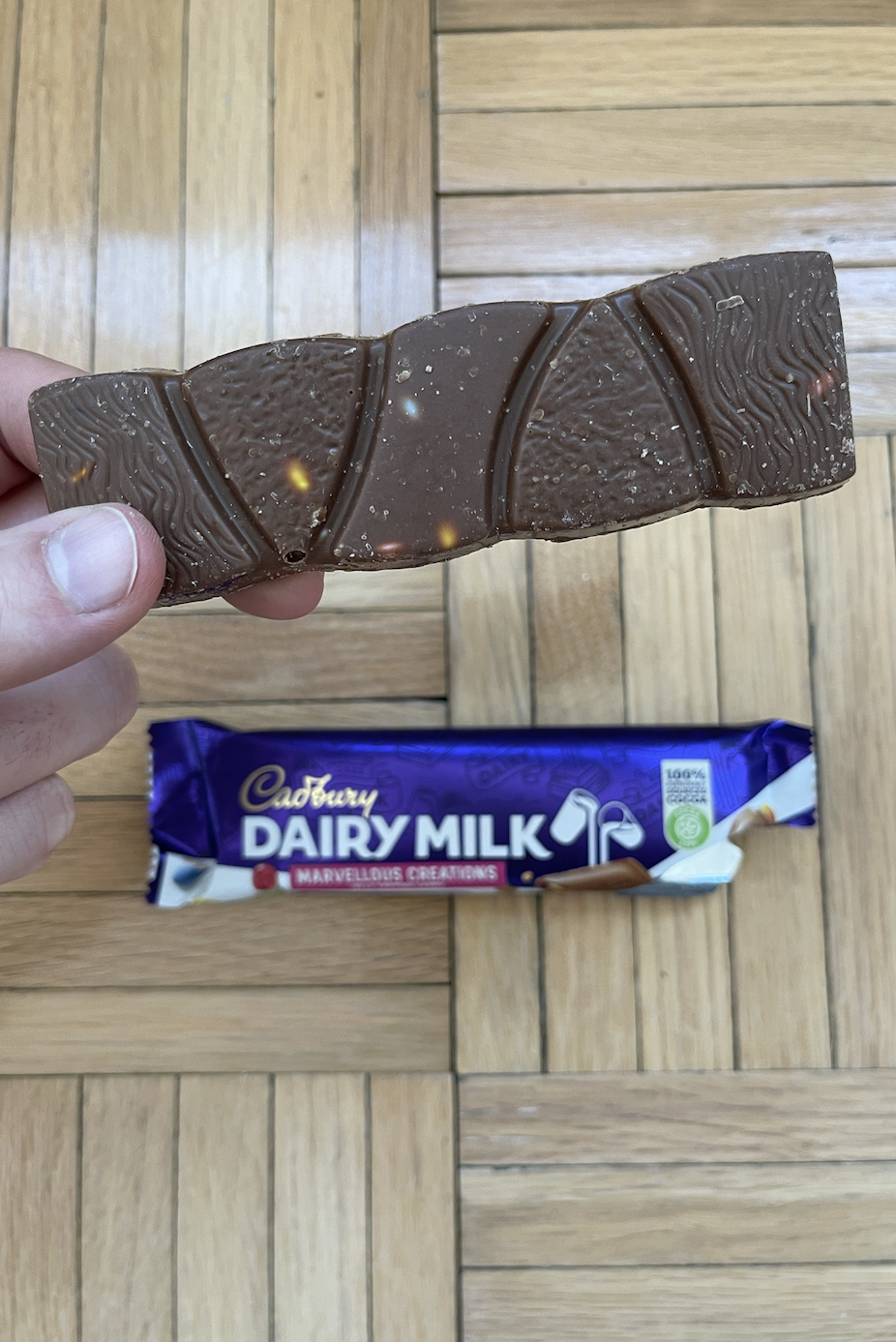 Hand holding a piece of Cadbury Dairy Milk chocolate above its wrapper on a wooden floor