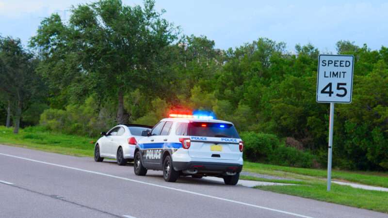 A traffic stop shows a police cruiser with flashing lights sitting behind a white car on the side of the road