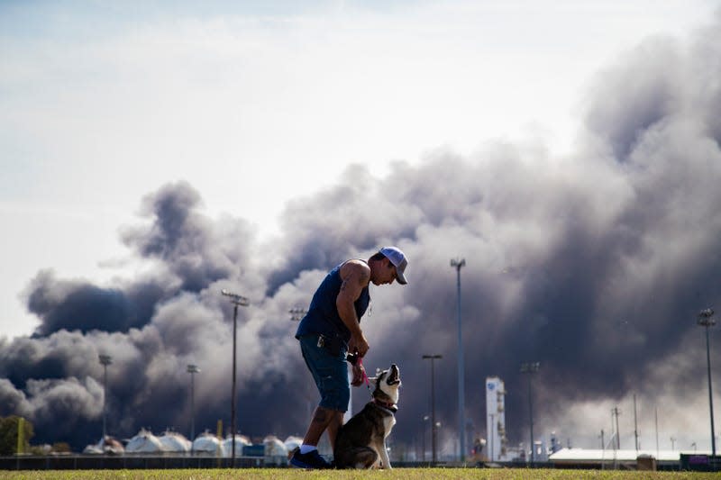 A man in shorts and a ballcap walks his dog in front of a massive plume of black smoke
