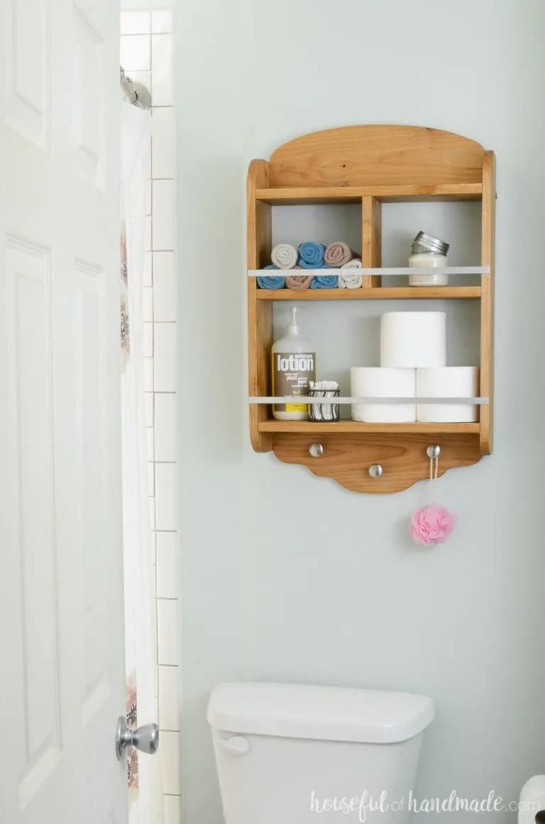 wood shelving above the toilet in a bathroom with toilet paper, lotion, a cup with q tip and rolled washcloths on it, there are silver pegs at the bottom and a pink bath