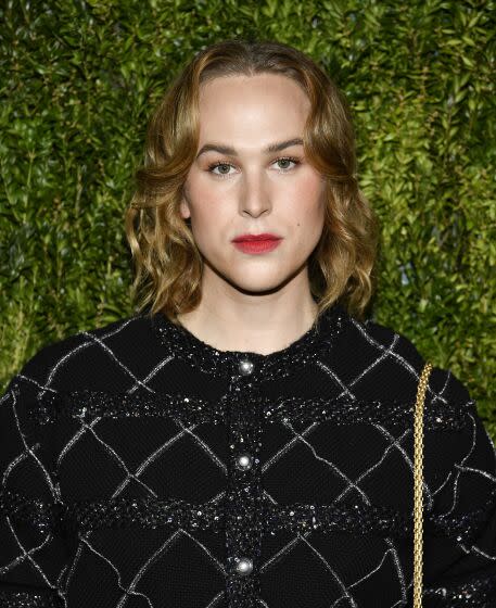 Tommy Dorfman wearing red lipstick and a sparkling black top poses in front of a greenery backdrop