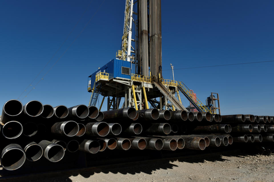 More than 50 large-diameter pipes with threaded ends stacked against a blue sky and drilling equipment.