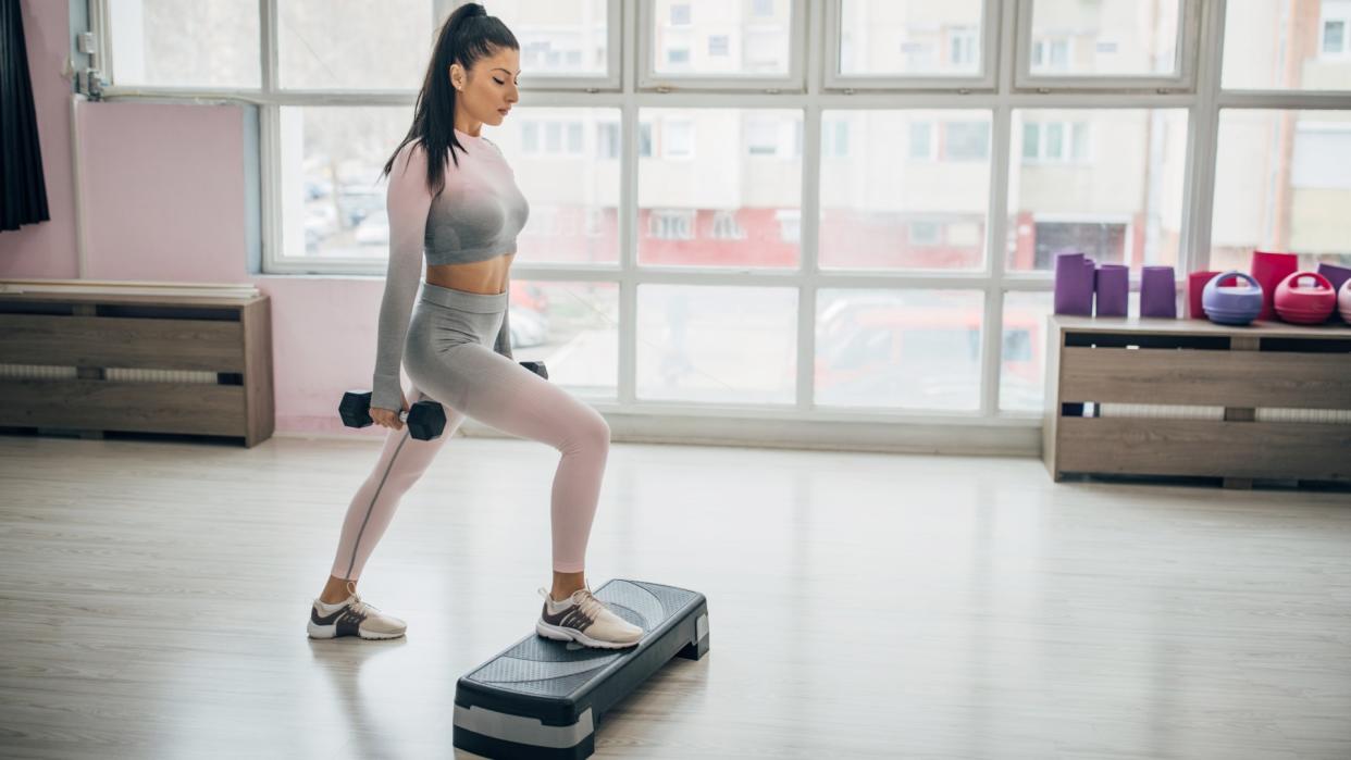  Woman holding light dumbbells stepping up onto a step during walking workout with weights. 