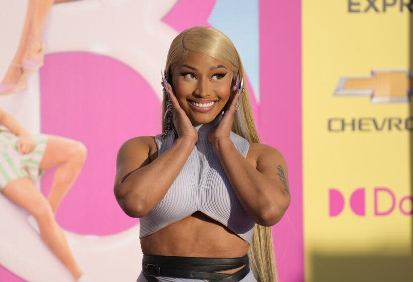 Nicki Minaj in a matching blue-gray top and skirt holding her hands to her face and smiling in front of a Barbie poster