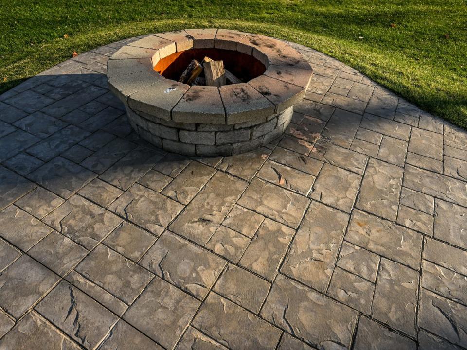 A fire pit with wood in it sitting along a patio with stamped concrete and grass around the edges of the area