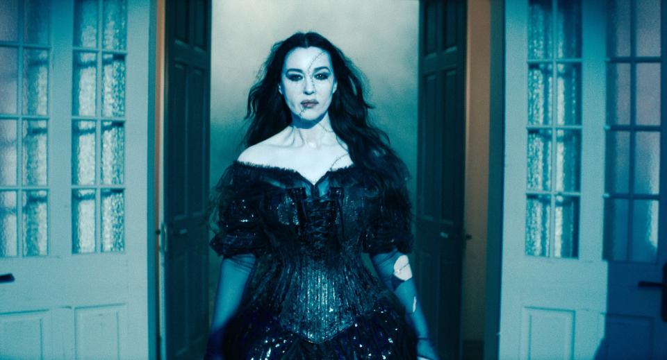 Monica Bellucci stands confidently between open doors in a black gothic-style dress with sequins, portraying a dark, mysterious character