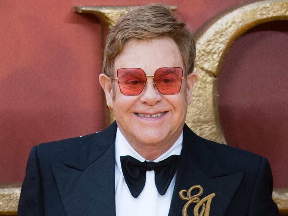 Elton John, wearing pink-tinted sunglasses and a velvet tux with floral detailing and an embroidered EJ, poses on the red carpet.