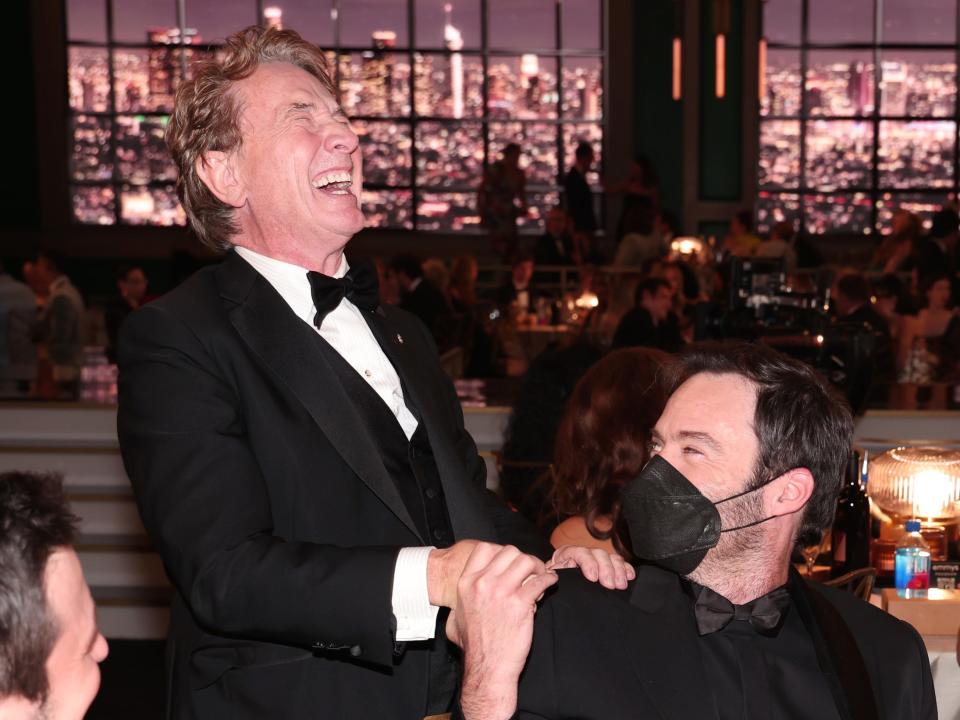 martin short resting his hand on bill hader's shoulder, where bill is grasping it. bill, who is wearing a mask, and martin are both laughing