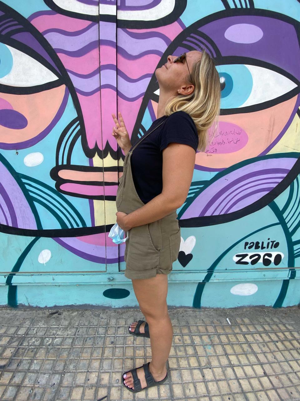 A woman who is pregnant standing sideways holding her stomach and giving the peace sign in front of a colorful mural.