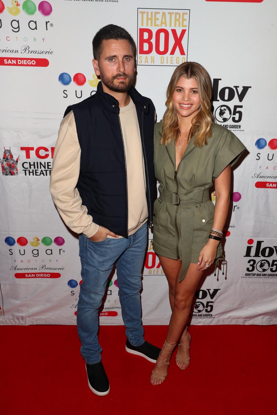 Scott Disick stuns in arm-less jacket and long-sleeve with sneakers beside Sofia Richie who is dressed in a green low-cut short-sleeve dress.