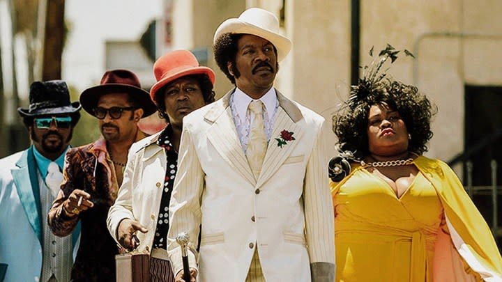 Eddie Murphy with a white suit, top hat, and cane, others following behind him in a scene from My Name is Dolemite.