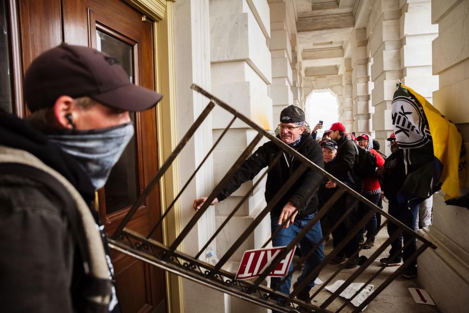 A rioter uses railing to bash a door at the Capitol as others stand by filming his attempt to gain access to the building.