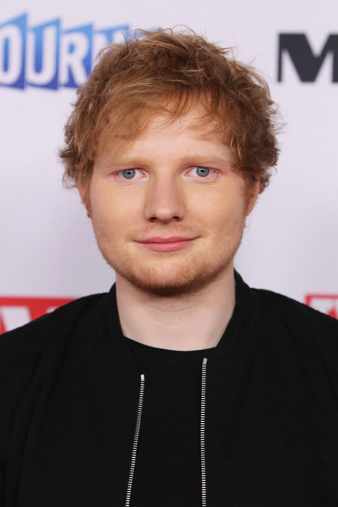 Ed Sheeran at an event, wearing a black jacket over a black shirt, against a backdrop with logos and text