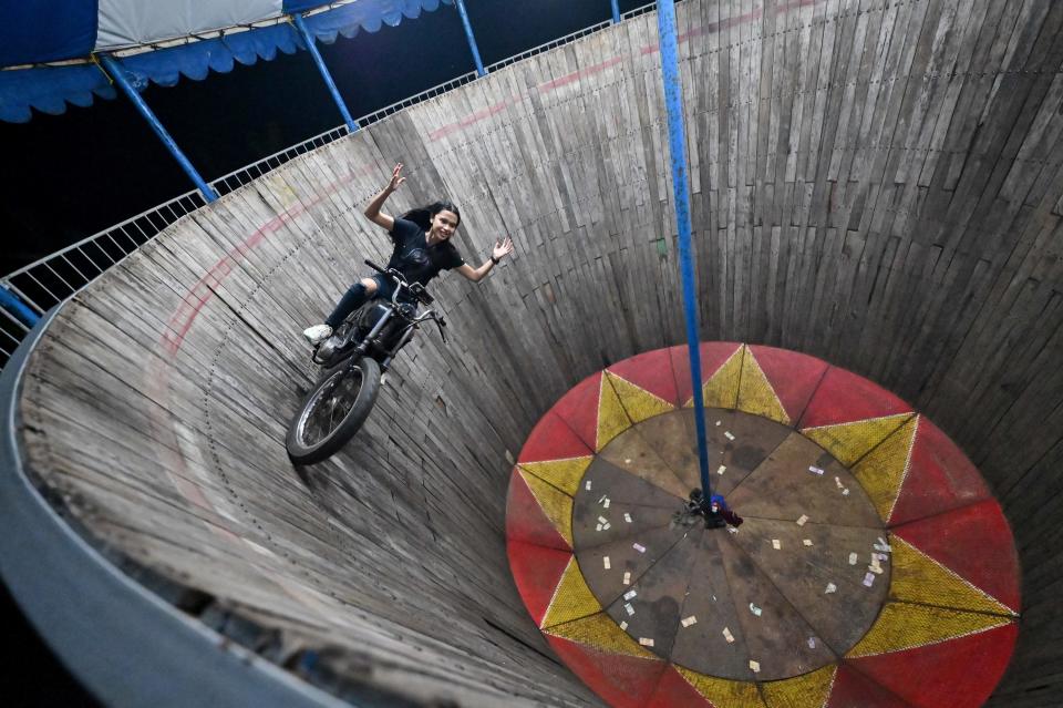 Karmila Purba, a young daredevil, rides the walls of the "Well Of Death." She is smiling and throwing her hands in the air, vertical to the ground on a motorcycle.