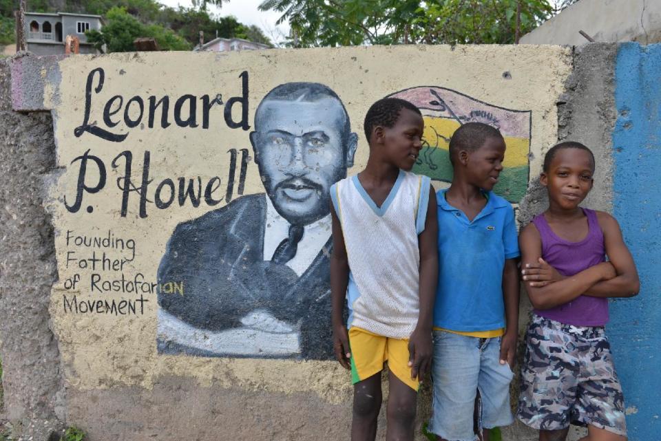 In this Jan. 4, 2014 photo, students pose for a photo by a mural of Rastafarian founding father Leonard P. Howell in Tredegar Park, Jamaica. The town of Tredegar Park is below a hilltop known as “Pinnacle” where Rastafarians lay claim to what they say is a sacred site where Howell once led a flourishing community in the 1940s. Ruins where Howell's house once stood were declared a national heritage site by the government last year. (AP Photo/David McFadden)