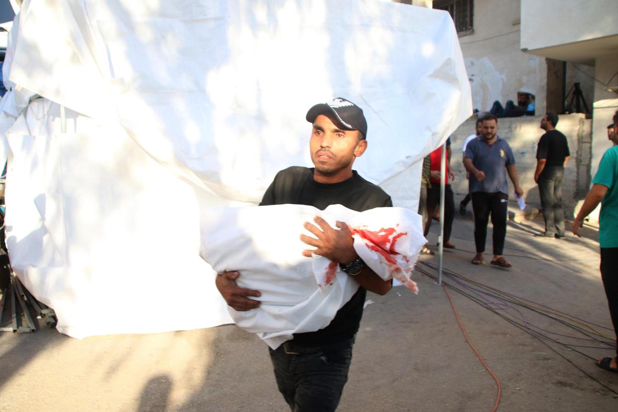 A man carrying the body of a child, draped in a white sheet that is bloodstained, walking through the street. His eyes look numb. 