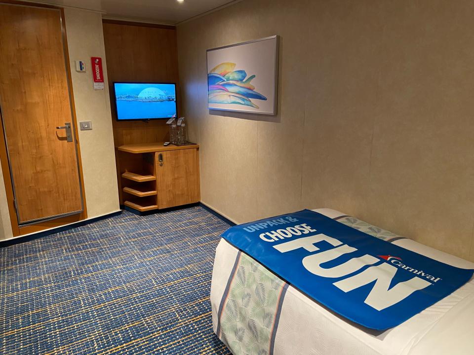 A cruise ship cabin with a bed with a blanket that says fun in caps and a small TV with a cabinet under it.