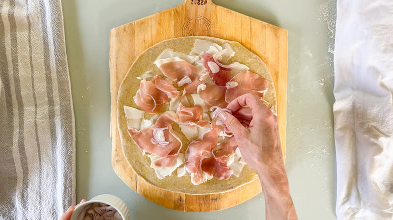 Putting sliced shallots on flatbread with shaved goat gouda and torn prosciutto on pizza peel