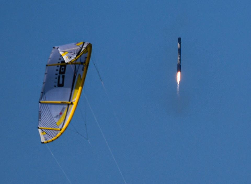 The Falcon 9 booster from Thursday's launch descends past the sail of a kiteboarder as it prepares to land at Cape Canaveral Space Force Station.