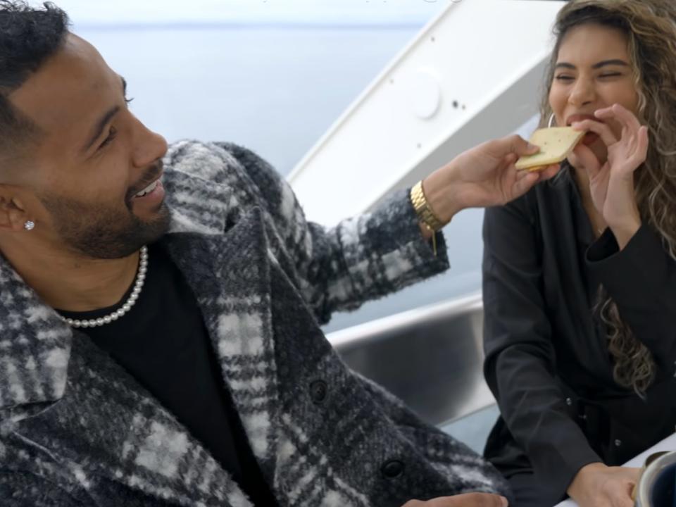 Josh, in a plaid coat, feeds Jackie, in a black shirt, a slice of cheese while sitting in a Ferris wheel compartment. They're both smiling, and a bottle of champagne sits on the table in front of them.