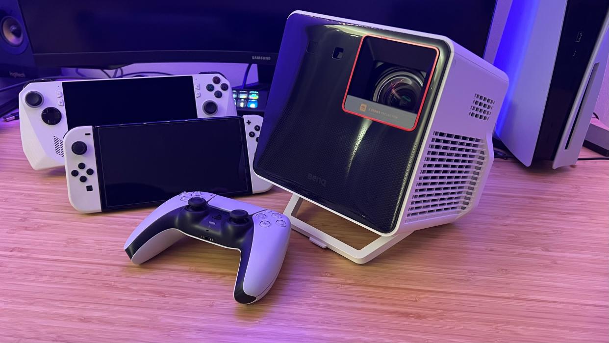  BenQ X300G projector on a wooden desk with Nintendo Switch, Asus ROG Ally, and PS5 controller. 