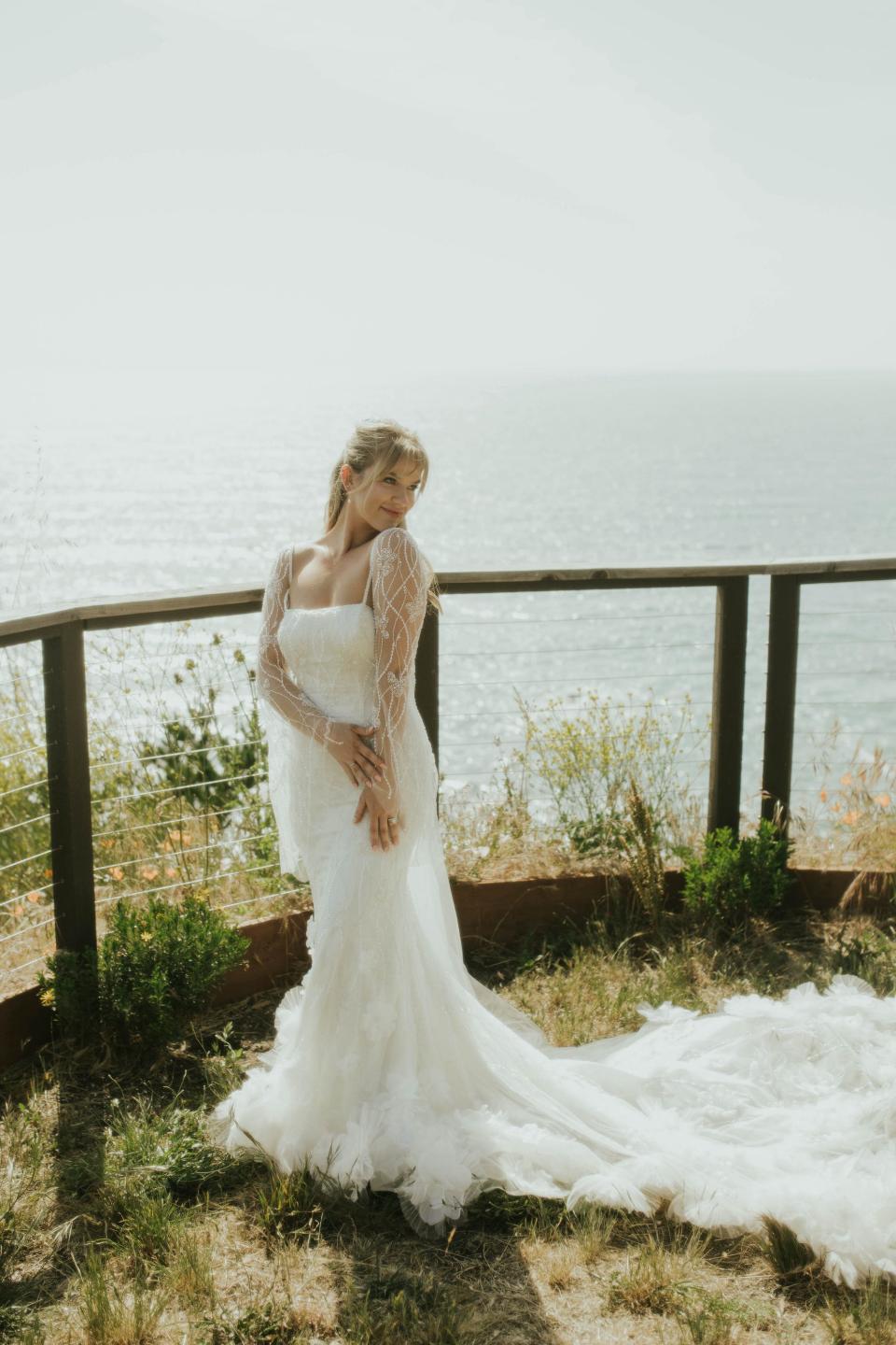 A bride stands in her wedding dress in front of a fence overlooking the ocean.