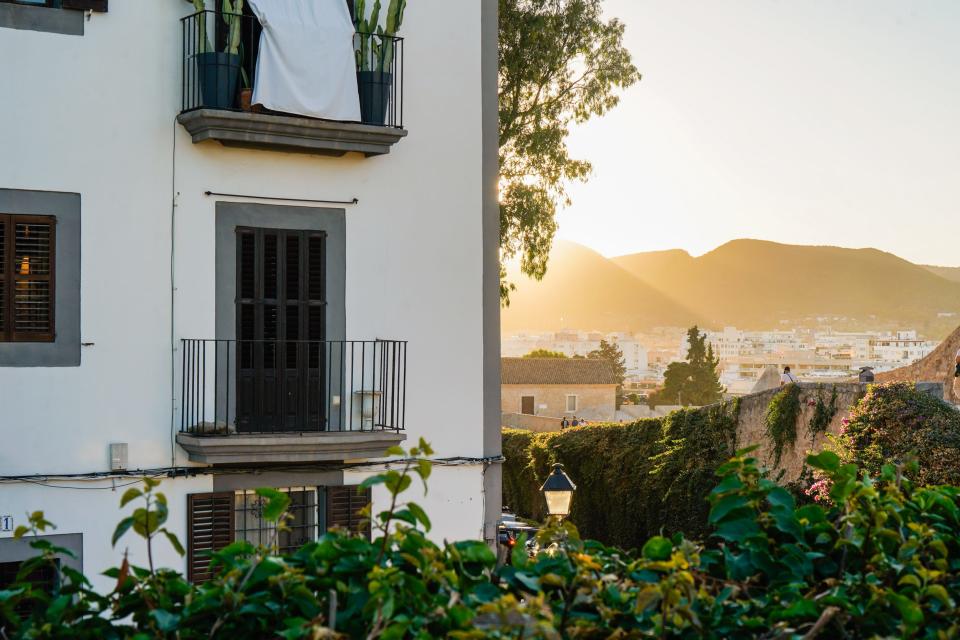 A white home in Ibiza with foliage in the foreground and the sun setting over mountains in the background