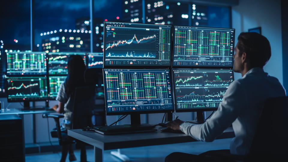 Hedge fund analysts review financial data in a dark room on several computer screens.